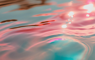 Primo piano ravvicinato e astratto di onde e increspature fluide sulla superficie dell'acqua, con riflessi di luce caldi nei toni del rosa antico, dell'ambra e tocchi di azzurro e teal sul fondo